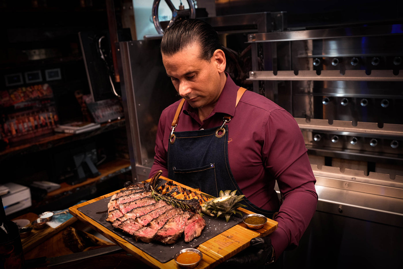 Man in a kitchen holding a tray with a large cut of meat and vegetables.